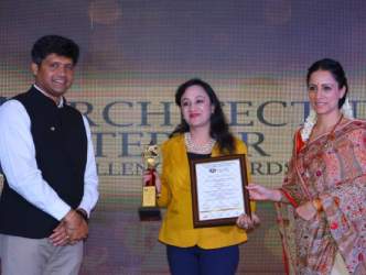 Woman receiving interior design award on stage, holding trophy and certificate with two presenters.