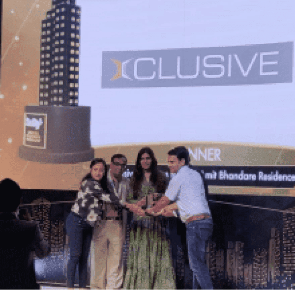 Group receiving award on stage with 'XCLUSIVE' logo and cityscape-themed backdrop.